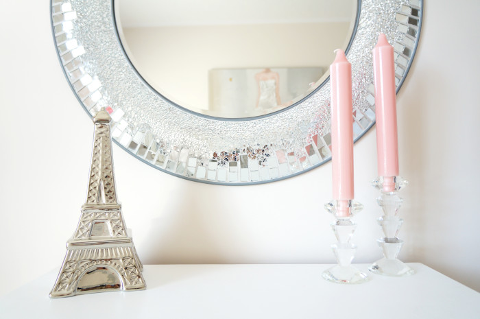Circular silver-framed mirror hung over a white surface topped with a silver Eiffel tower and two pink candlesticks