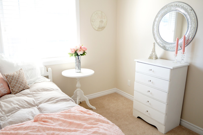 Bright room with pink flowers, a pink and white bed, a white dresser, and a circular silver-framed mirror