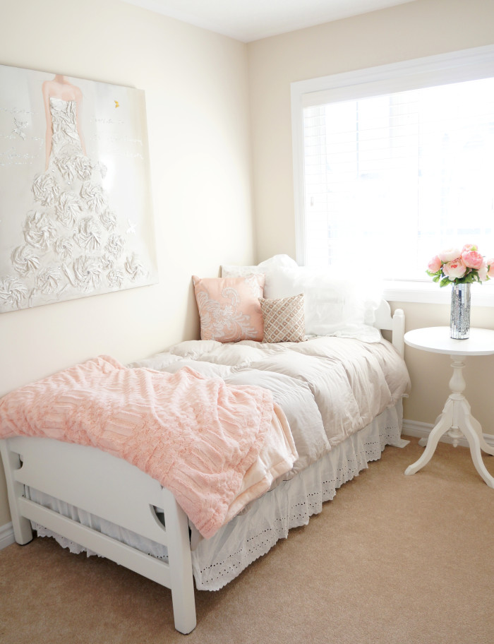 Pink and white bed in the corner of a room with a white nightstand, pink flowers, and a wall hanging with a textured wedding dress