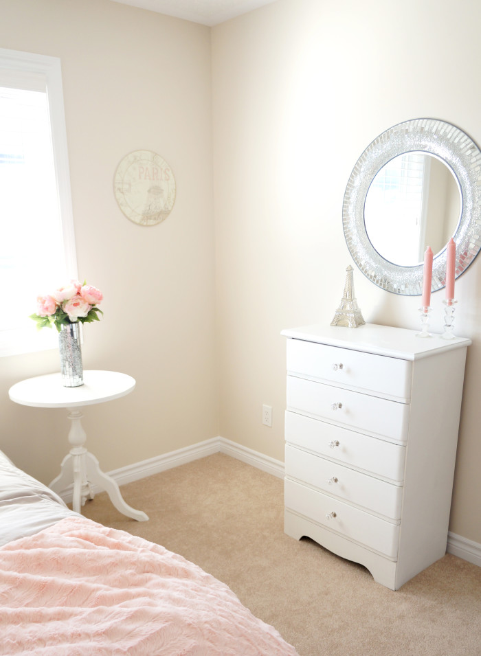 Corner of a bright room with pink flowers, a silver-framed circular mirror, and a white dresser with pink candles and a mini Eiffel tower