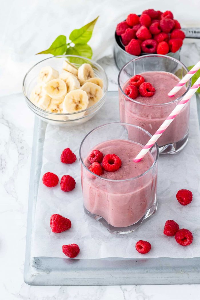 raspberry and banana smoothie in glasses with bowl of bananas and raspberries