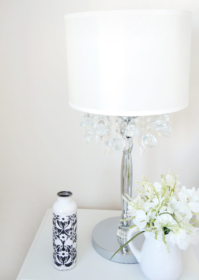 Black and white water bottle on a nightstand next to a lamp and a vase of white flowers