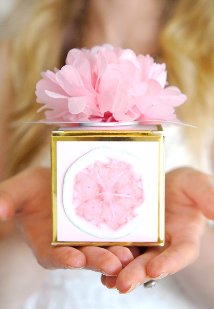 Hands holding a golden box topped with a pink flower and decorated with a picture of a watermelon