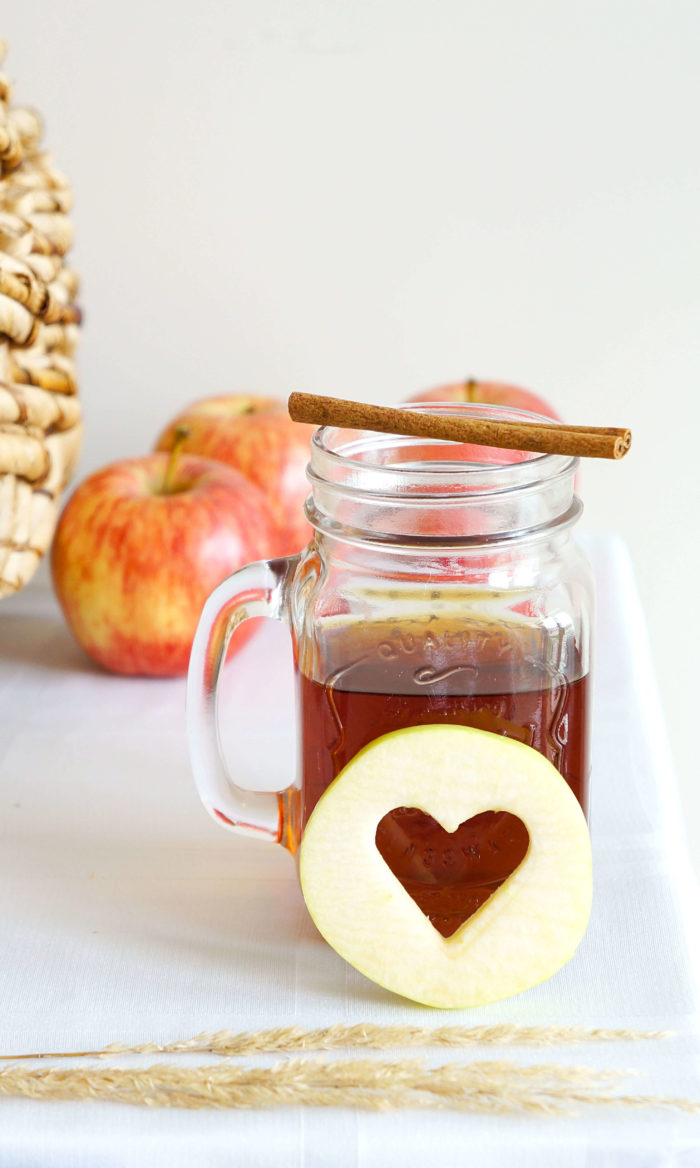 Mason jar topped with a cinnamon stick next to an apple with a heart-shaped piece missing