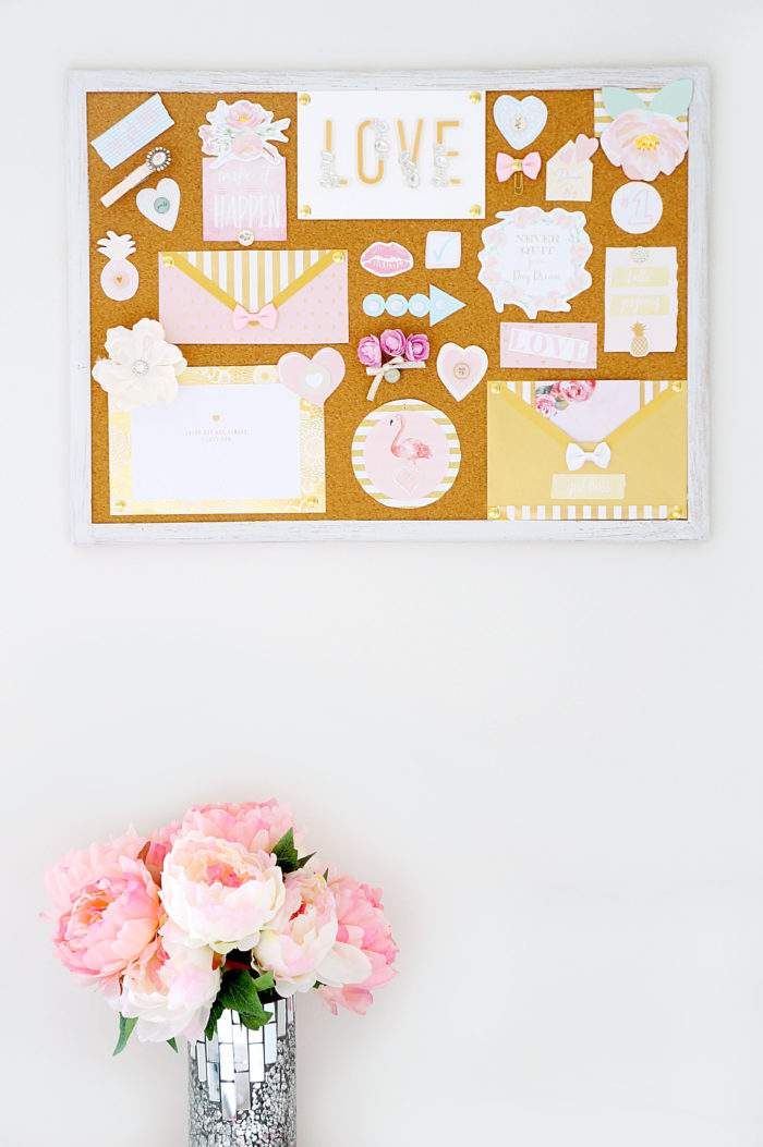 Cork board full of pastel decorations and papers hanging above a silvery vase of pink flowers