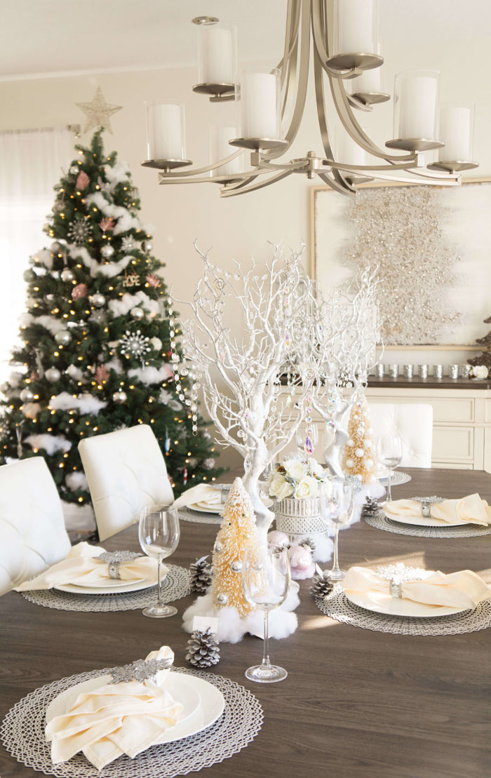 Table set with silver, ivory, and sparkly Christmas decorations with a green Christmas tree in the background