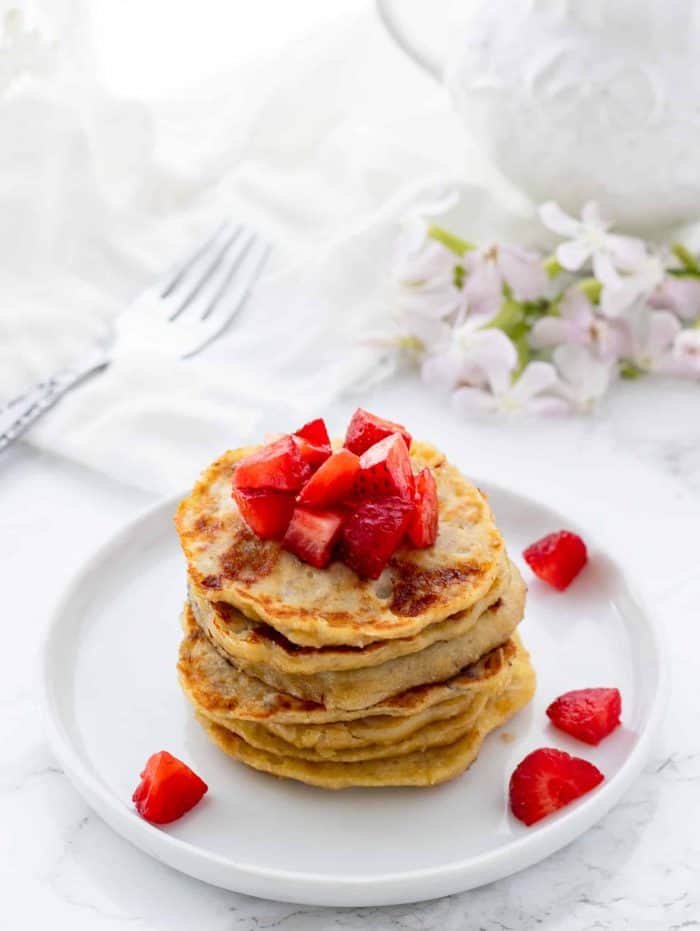 stack of pancakes topped with diced strawberries on white plate with fork and flowers in background