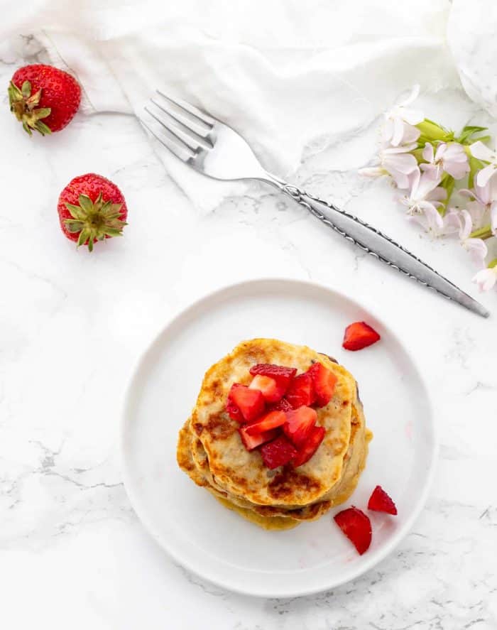 stack of mini pancakes topped with diced strawberries on a white plate surrounded by a fork and fresh strawberries