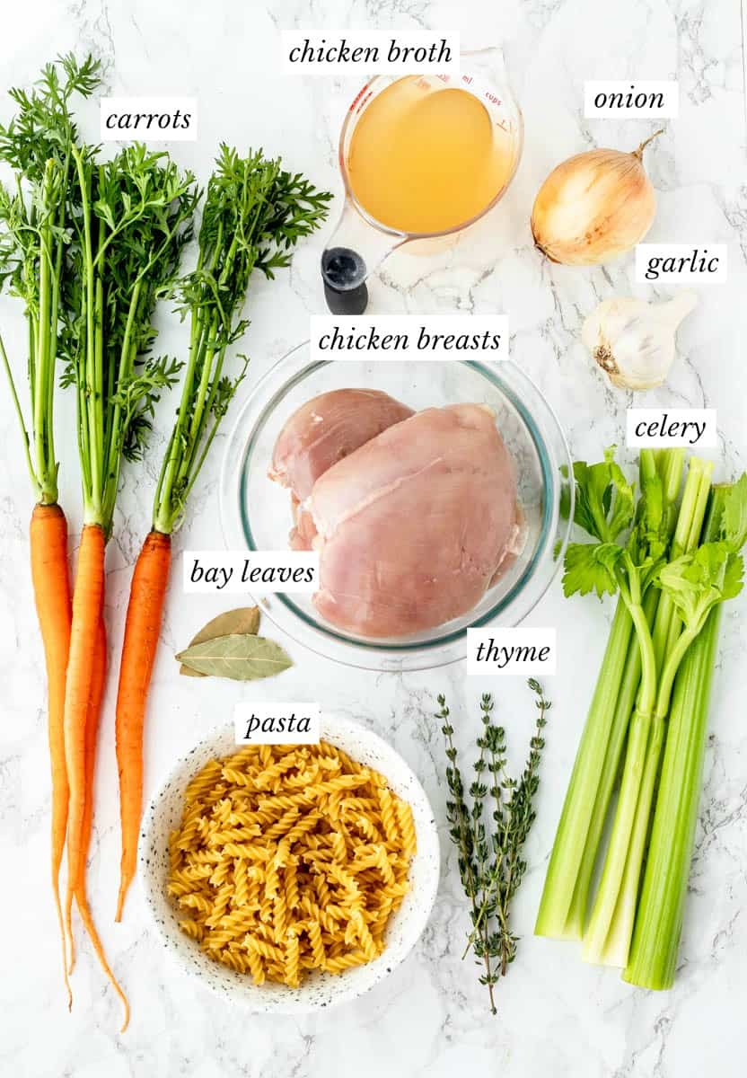 Ingredients required to make healthy chicken noodle soup with labels on a marble background.