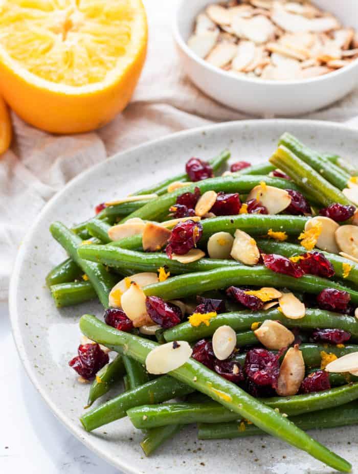 green beans with garlic, dried cranberries, almonds and orange zest on plate with orange and almonds in background