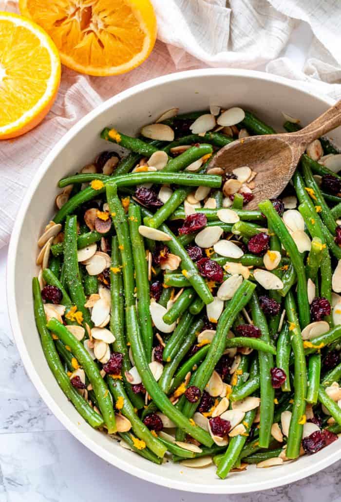 green beans with garlic, dried cranberries, almonds and orange zest in a large pan with a rustic wooden spoon next to fresh orange halves
