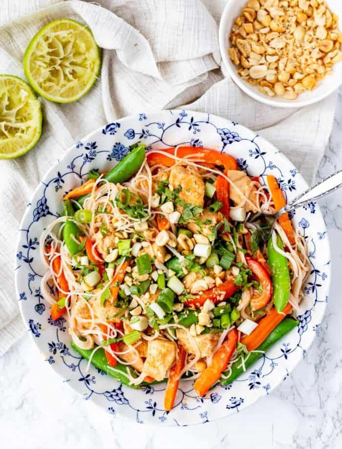 peanut chicken mixed with vegetables and rice noodles in a bowl with limes and peanuts