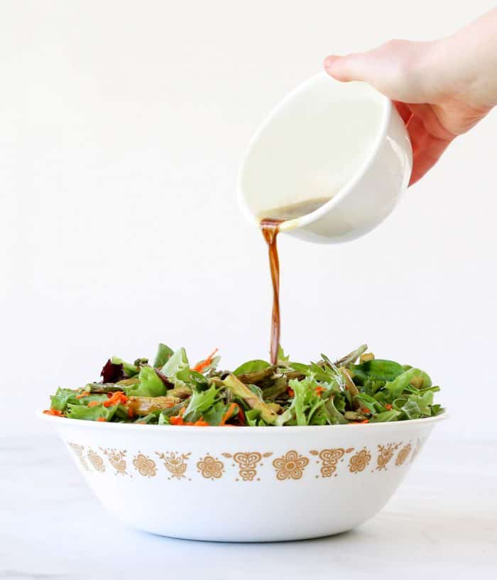 Pouring balsamic dressing over the salad in a white serving bowl