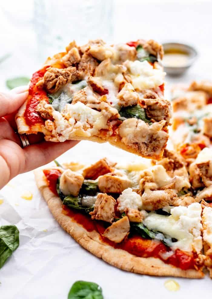 A hand holding a slice of chicken spinach pita pizza.