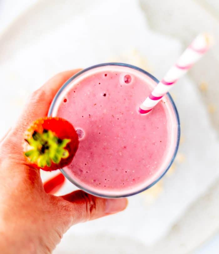 A hand holding glass filled with smoothie with a pink striped straw and strawberry