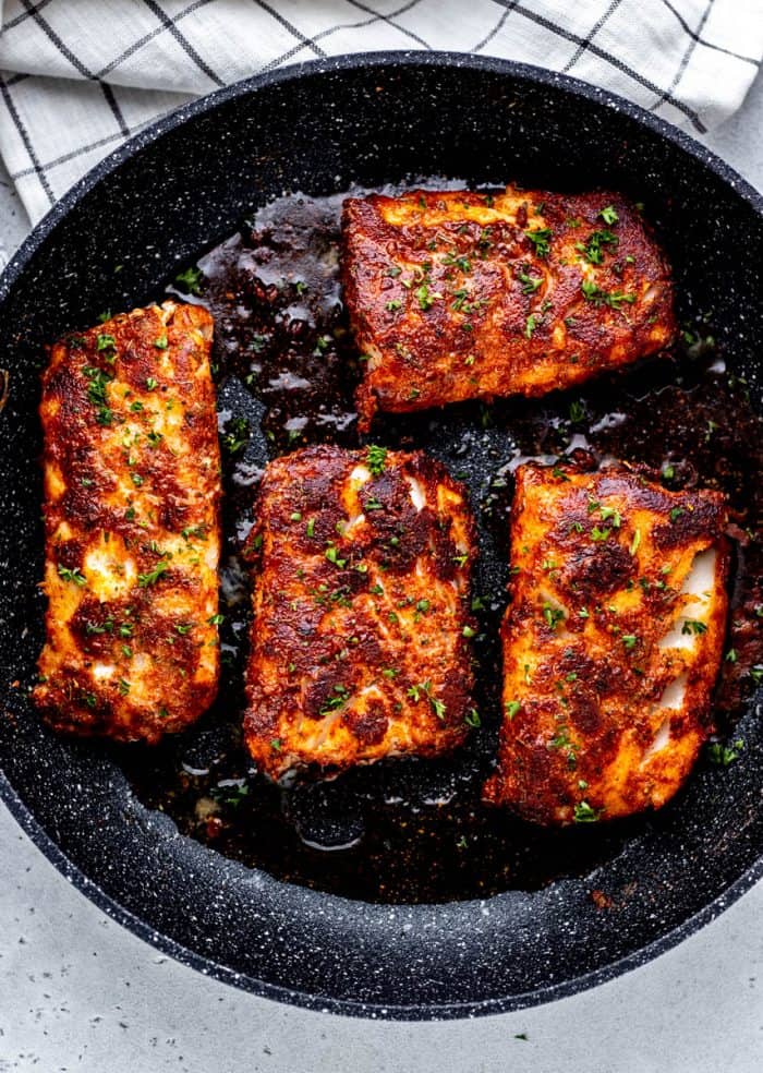 Four blackened cod fillets in a skillet garnished with fresh herbs.