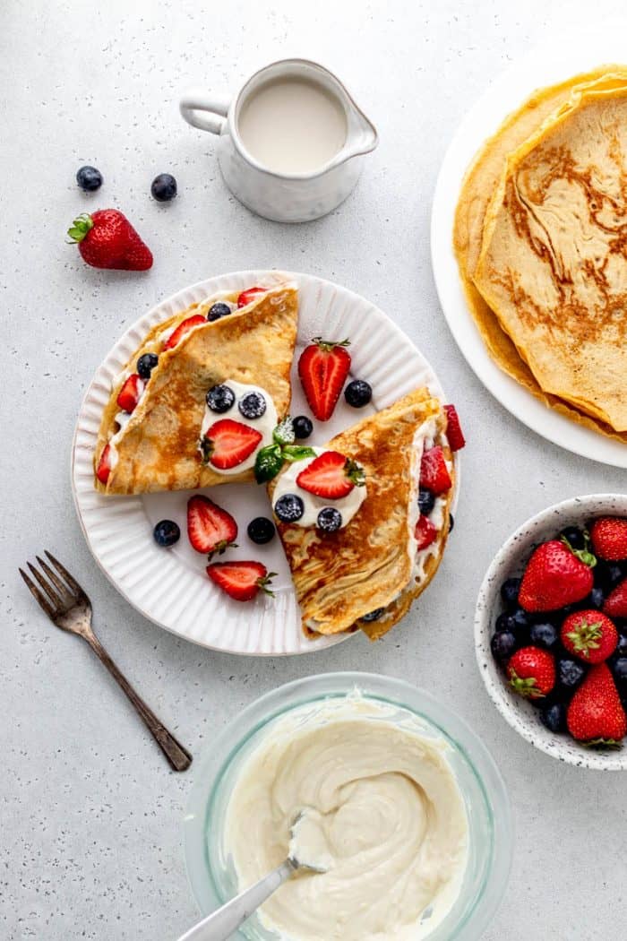 Two crepes on a plate next to bowls of fresh berries and cream cheese filling.