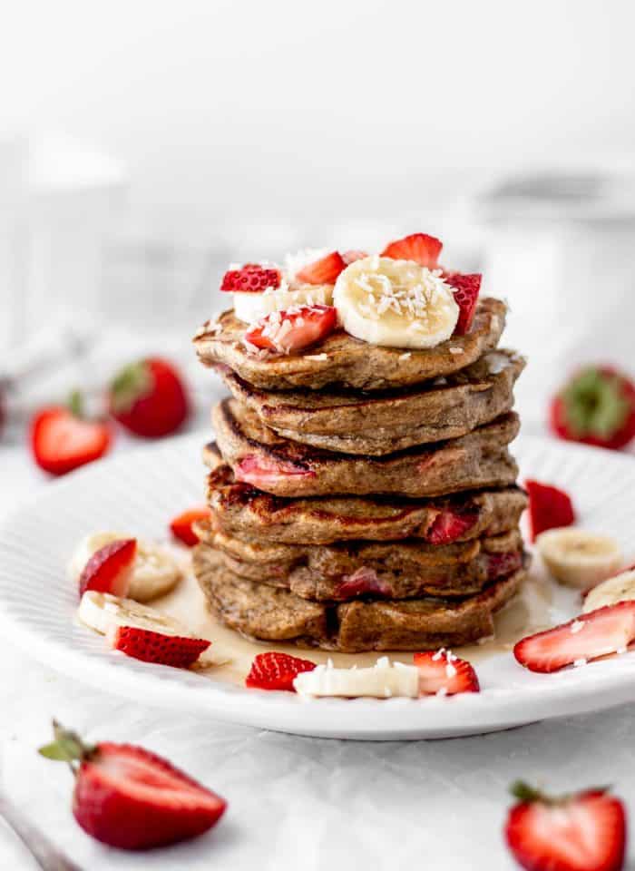 Stack of strawberry banana pancakes on a white plate topped with fresh fruit.