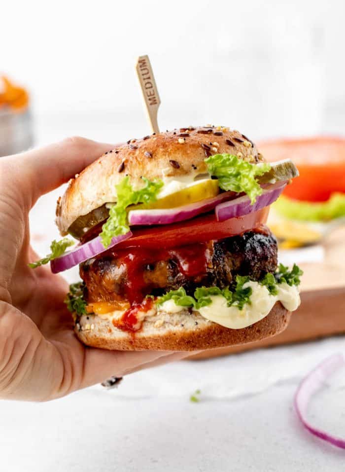 A hand holding a beef burger made with a seedy bun, lettuce, mayonnaise, pickles, red onion slices, a tomato slice, and ketchup.