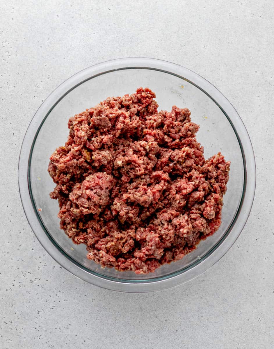 Raw ground beef in a large glass bowl mixed with other burger ingredients.