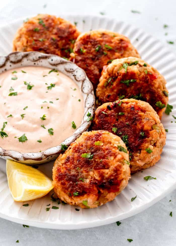 A close up image of fried salmon patties on a white plate around a bowl of creamy dipping sauce with a lemon wedge on the side.