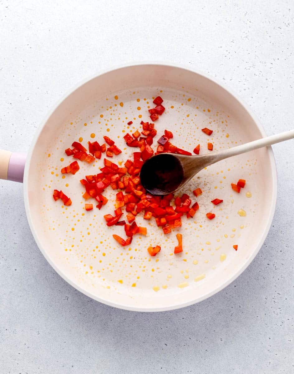 A skillet with chopped red pepper being stirred with a wooden spoon.