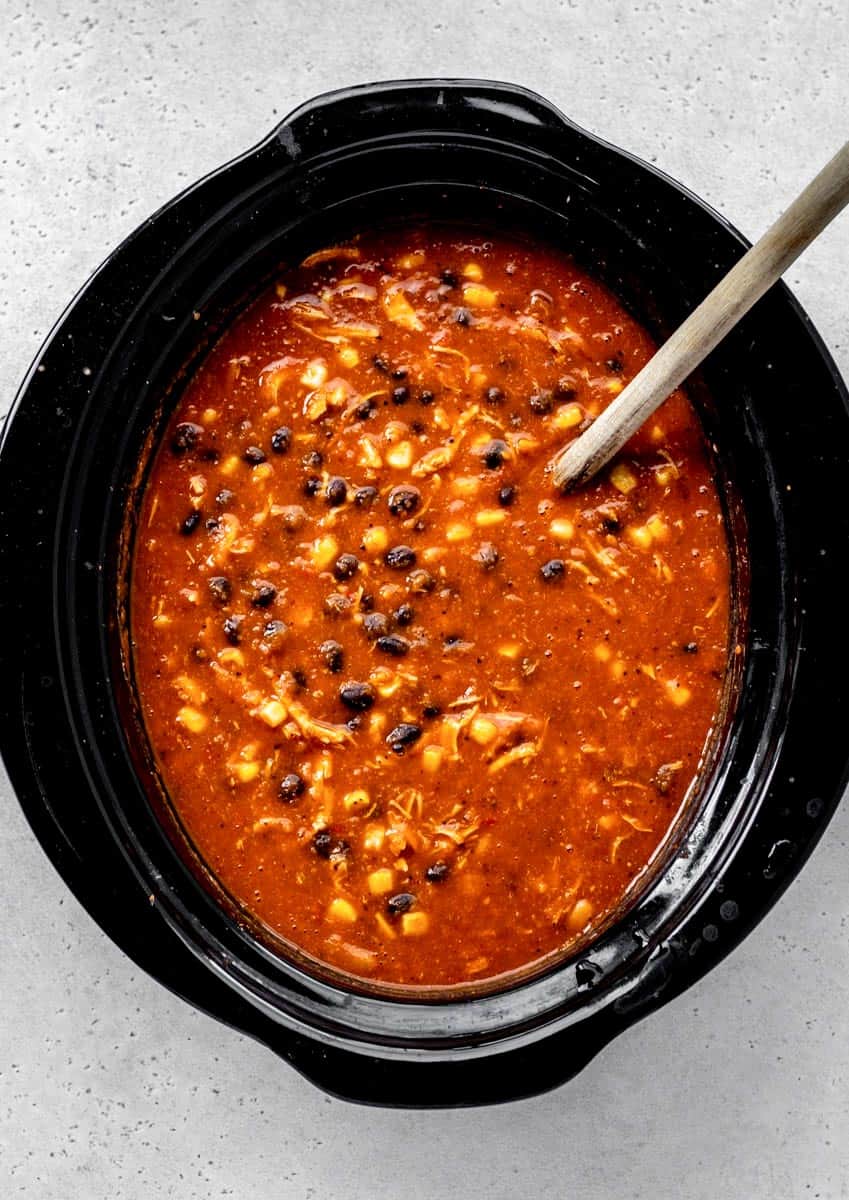 The creamy base in a slow cooker with chicken, black beans, and corn being stirred with a wooden spoon.