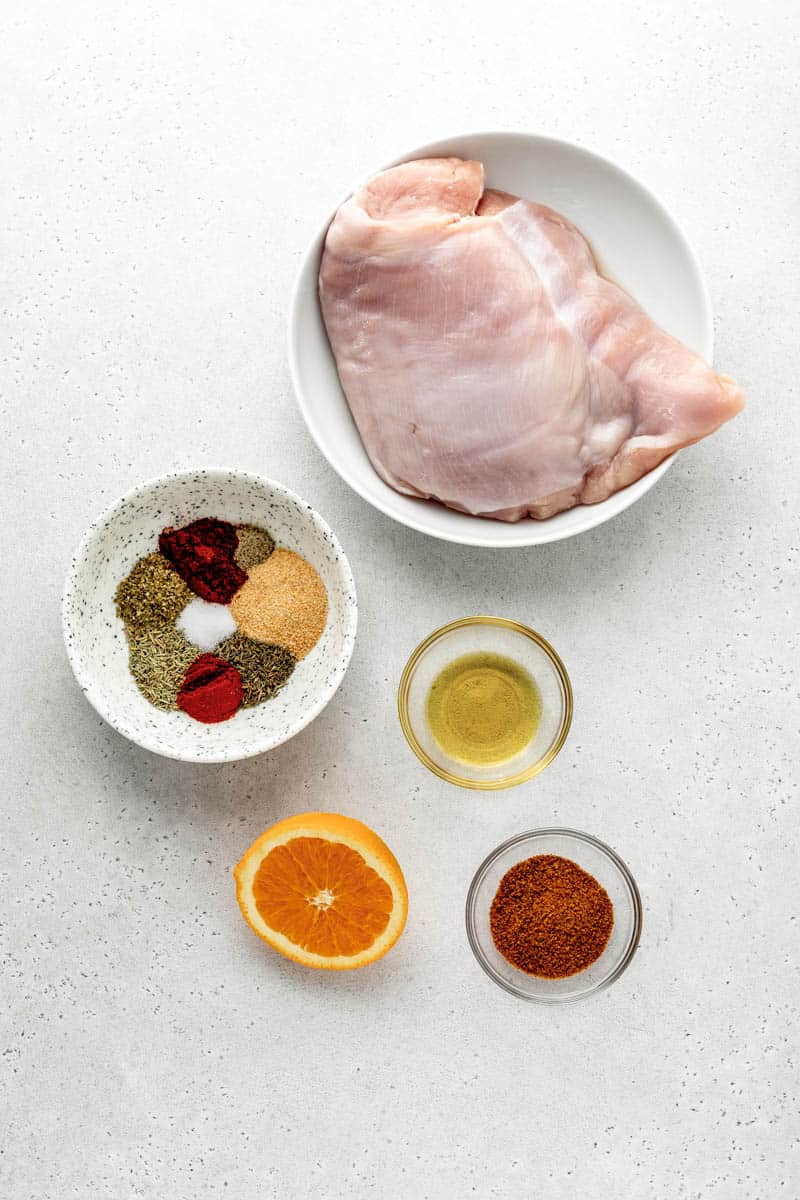 Overhead image of small bowls full of boneless turkey breast, spices, olive oil, coconut sugar, and half an orange.