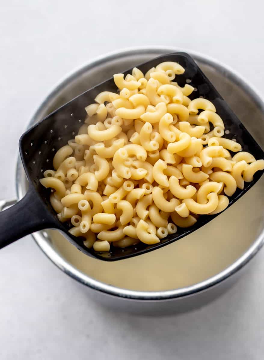 A closeup shot of cooked macaroni noodles on a scoop over a pot.