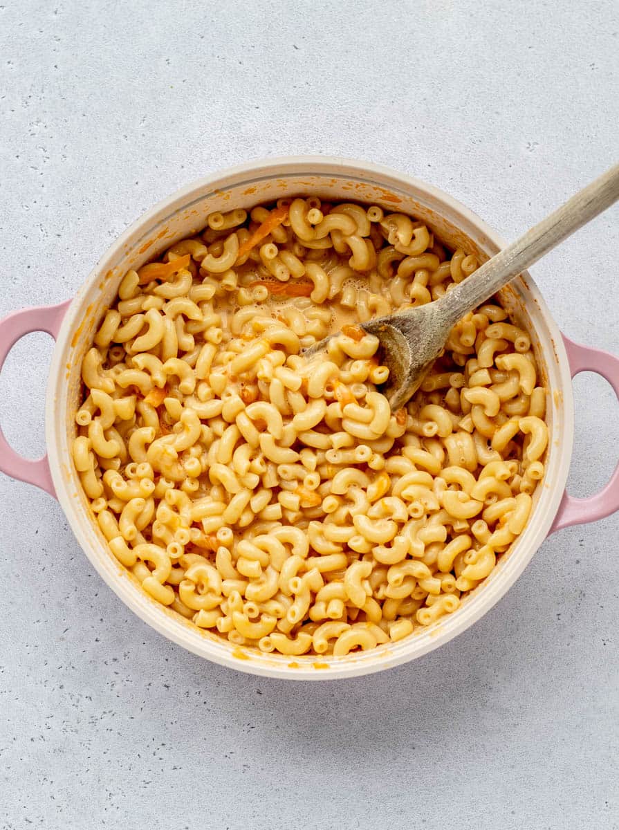 A top down view of a pot of evaporated milk mac and cheese. A wooden spoon is stirring sharp cheddar cheese into the pasta.
