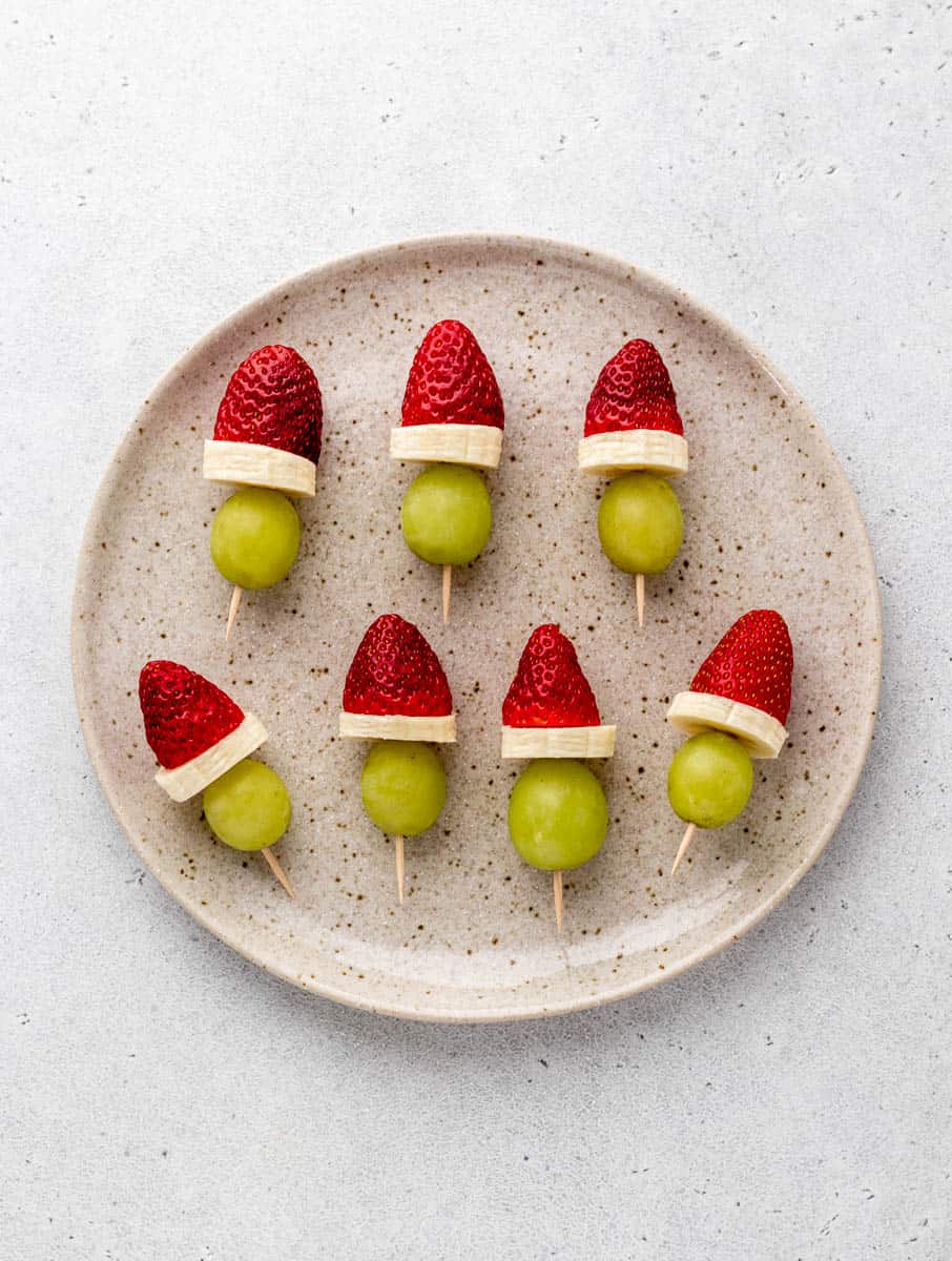 Seven grinch fruit kabobs on a speckled plate.