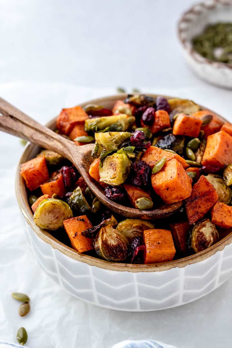 A wooden spoon in a serving bowl holding Maple roasted brussel sprouts and sweet potatoes with dried cranberries and pumpkin seeds.