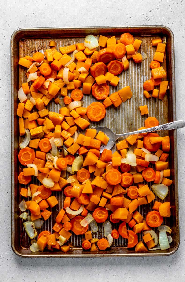 Chopped butternut squash, carrots, and onions in a single layer on a baking sheet being flipped with a spatula.