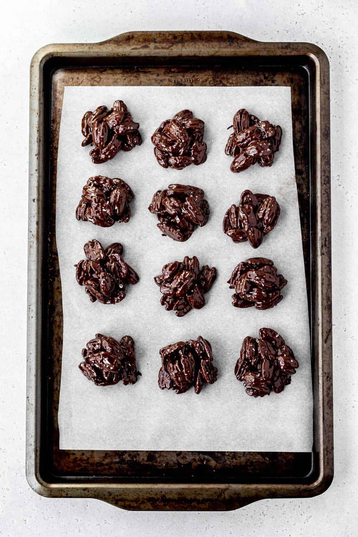 Freshly dipped dark chocolate pecan clusters on a parchment lined baking sheet.
