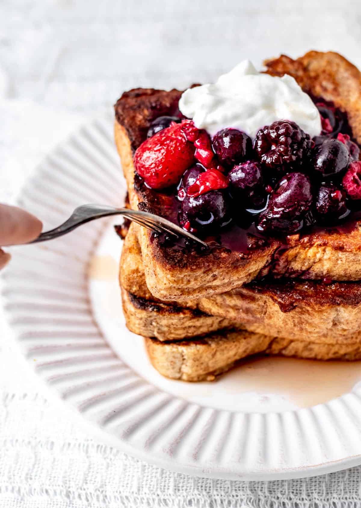 A fork poking into a stack of high protein French toast, topped with mixed berries, Greek yogurt and maple syrup.