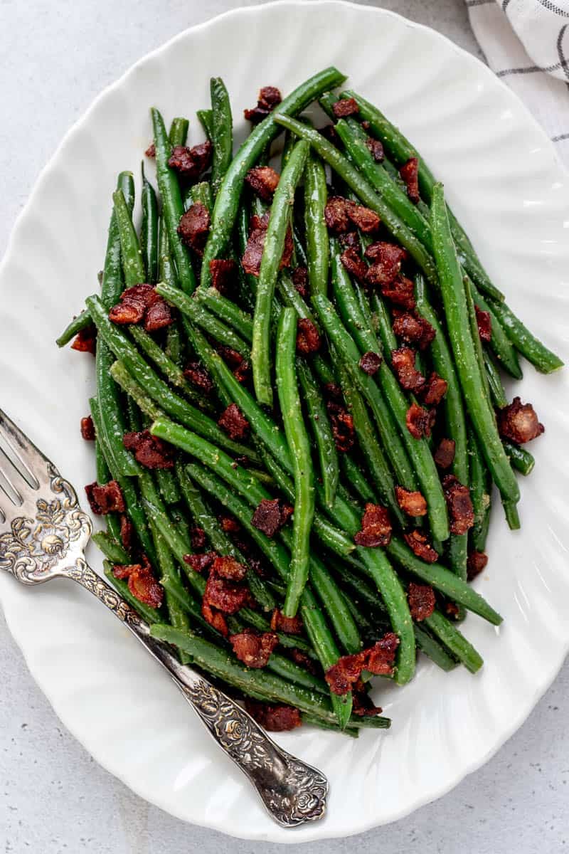 A white serving plate, with a fork on the side of seasoned green beans and bacon.