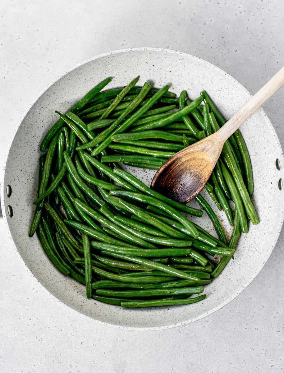 Green beans in a skillet getting sautéed with a wooden spoon.