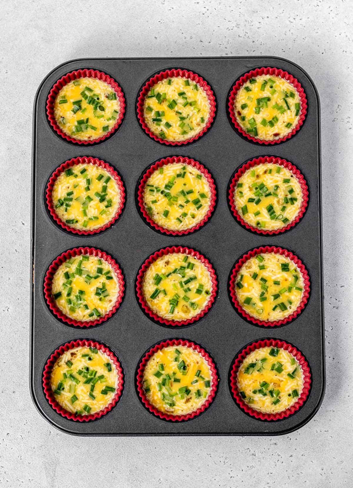 A muffin tin with 12 muffin cups filled with crustless mini quiches ready to be baked.