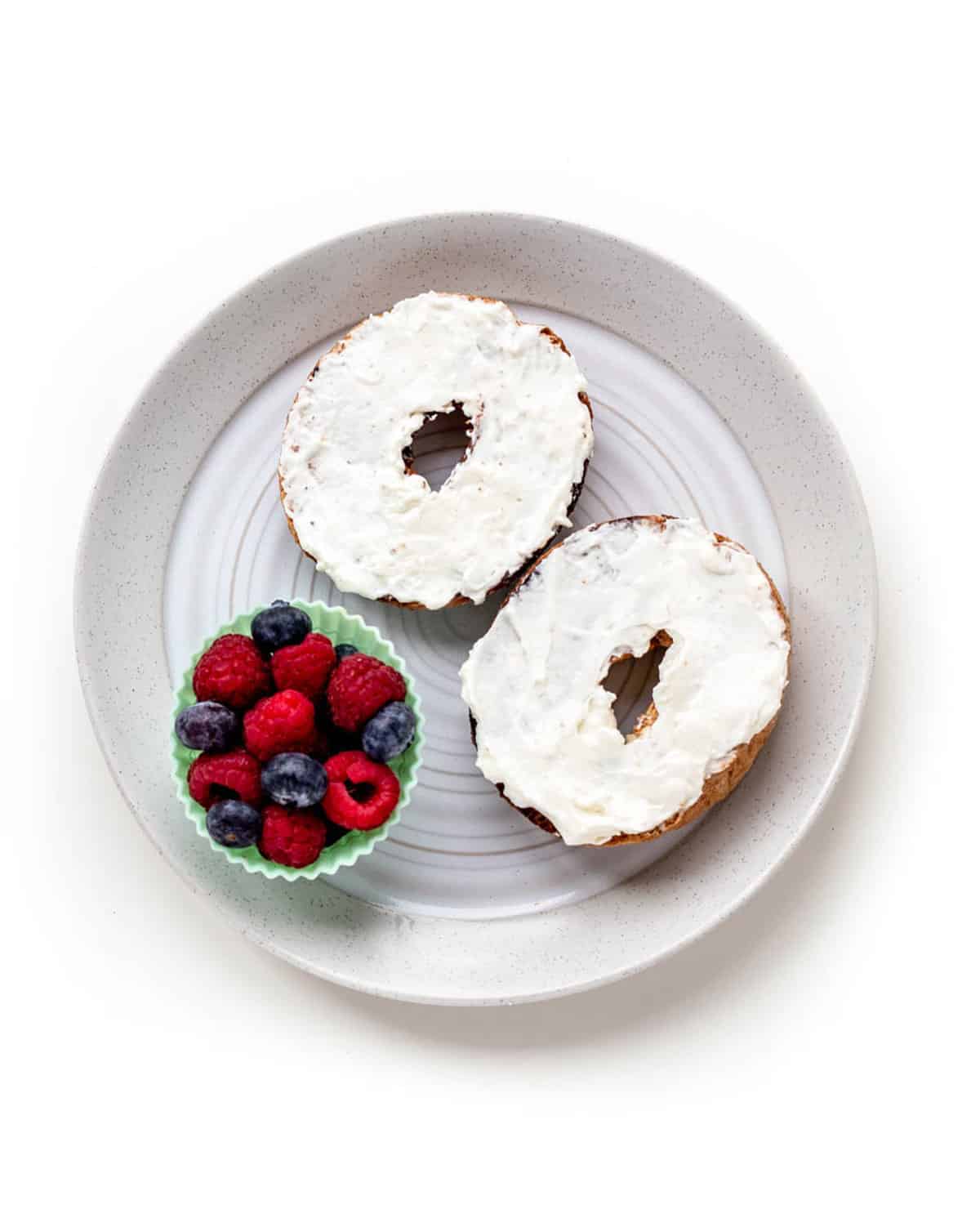 Bagel with cream cheese next to a cup of mixed berries on a plate.