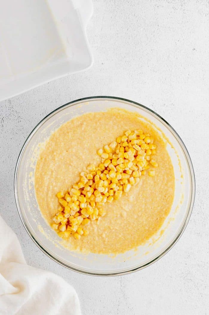 A glass bowl of cornbread batter with frozen corn kernels ready to be folded in.