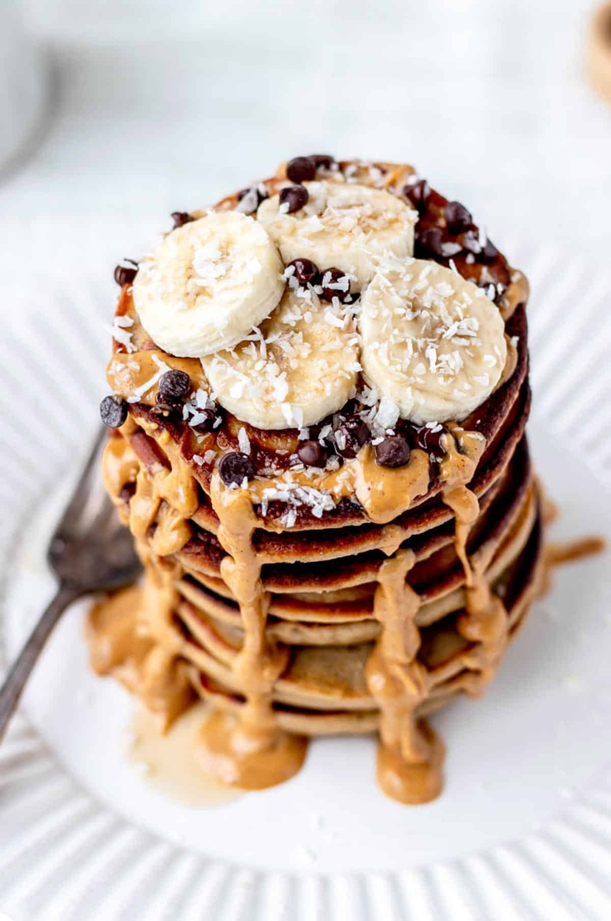 A stack of fluffy banana protein pancakes topped with banana slices, peanut butter, chocolate chips and coconut.