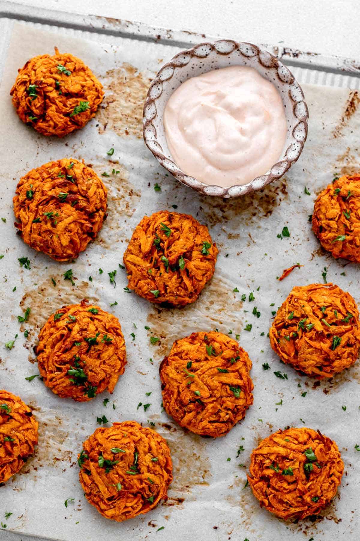 Overhead image of crispy sweet potato fritters on a parchment-lined baking sheet next to a bowl of dip.