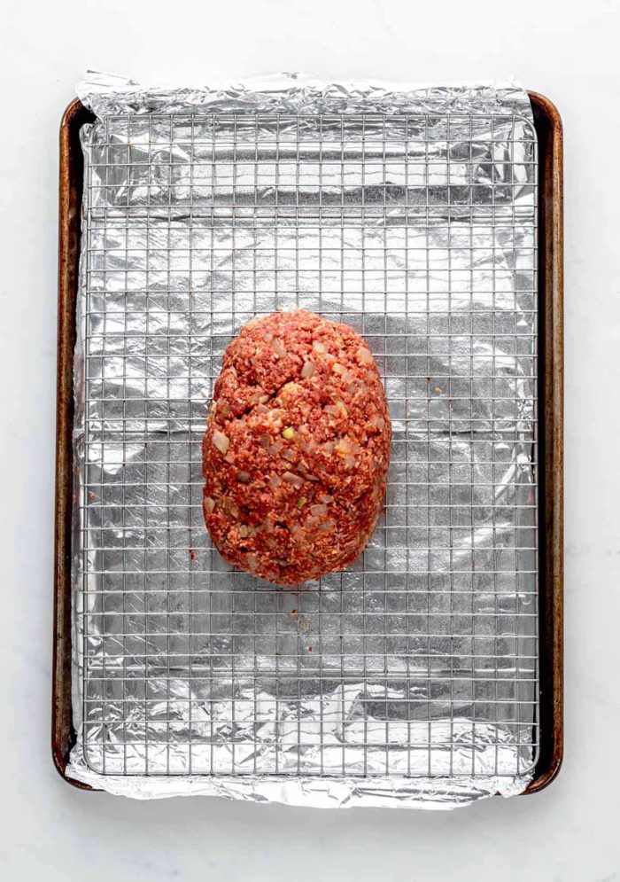 An aluminum foil-lined baking sheet, with a wire rack, with a one-pound meatloaf ready to be baked on it.