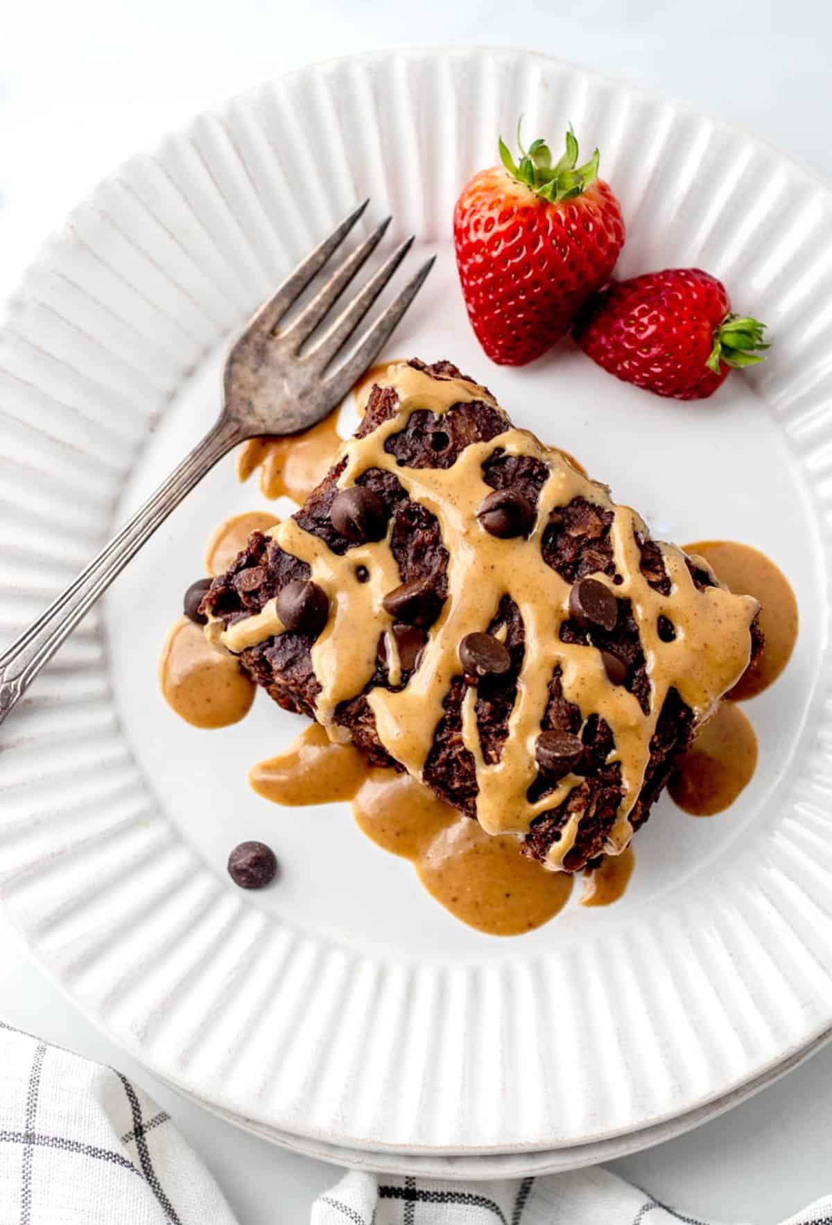An overhead image of chocolate baked oats on a plate topped with peanut butter next to two strawberries.